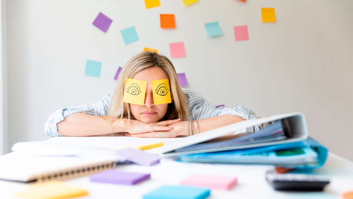 Frau am Schreibtisch, die mit dem Kopf auf den Armen liegt und bunte Post-it-Klebchen auf den Augen hat.