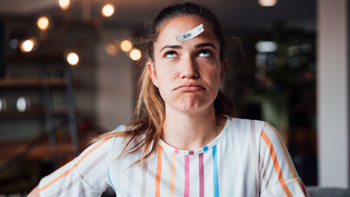 Eine Frau mit bunt gestreiftem T-Shirt, die in einem Zimmer sitzt und ein Post-it mit dem Schriftzug "To-do" auf der Stirn kleben hat