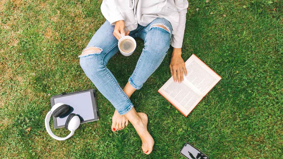 Bild aus Vogelperspektive von einer Frau, die auf einer Wiese sitzt. Sie hat einen Kaffee und ein Buch in der Hand und neben sich ein Tablet mit Kopfhörern und ein Smartphone liegen.