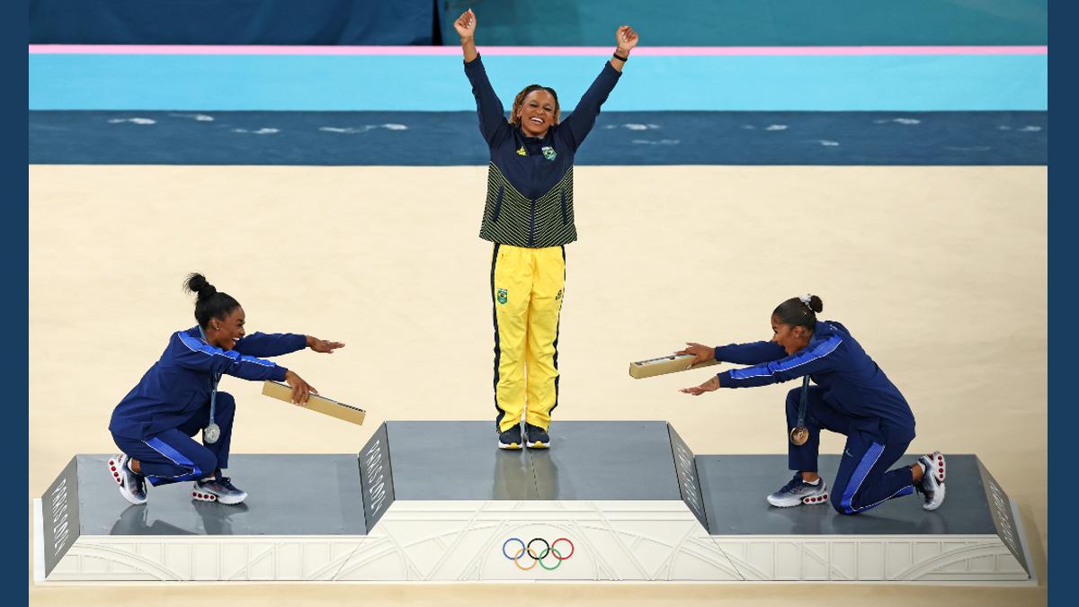 Das Bild zeigt die Siegerehrung der Bodenturnerinnen bei den Olympischen Spielen 2024. Siegerin ist Rebeca Andrade, Simone Biles (Silber) und die Drittplatzierte verneigen sich auf dem Siegerinnenpodest vor der Gold-Gewinnerin.