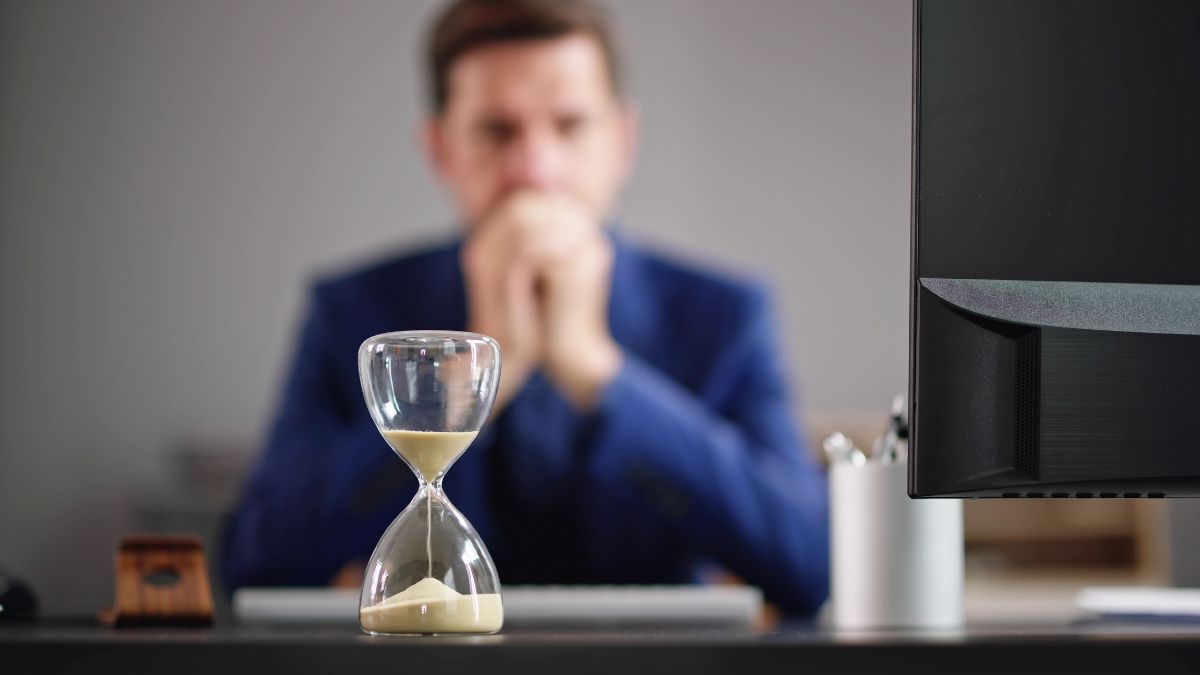 Eine Sanduhr steht auf eine Büro-Arbeitstisch, im Hintergrund ist etwas verschwommen ein Mann zu sehen