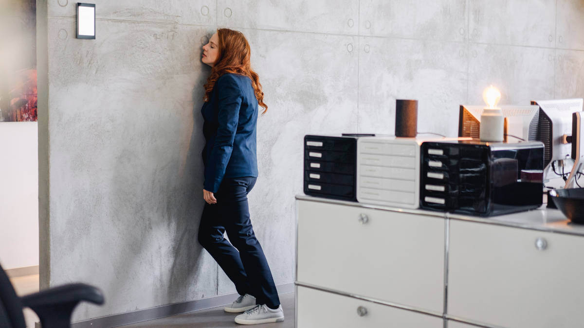 Eine rothaarige Frau mit blauer Kleidung, die in einem Büro mit dem Gesicht bei geschlossenen Augen an einer Wand lehnt.